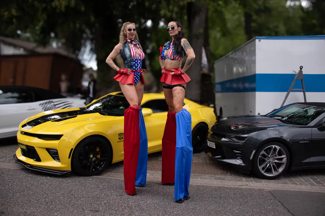 Duo d'échassières en costumes aux couleurs américaines, devant des voitures de sport.
